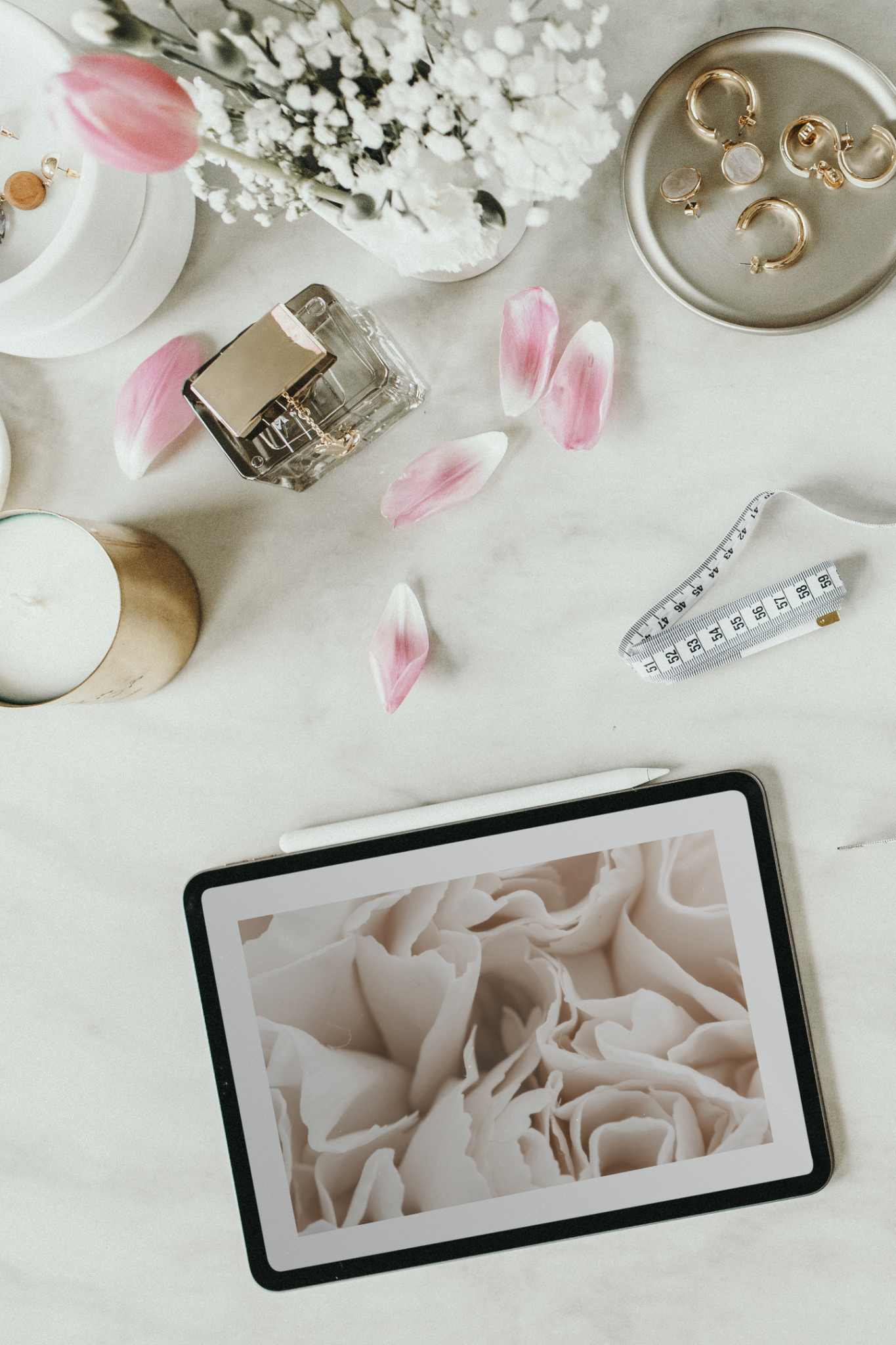 Aesthetic photo of iPad, flower petals and gold jewelry laid out on marble table.